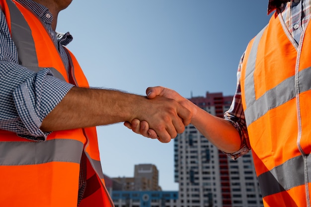 Des hommes vêtus de gilets de travail orange se serrent la main sur le chantier dans le contexte d'un immeuble de plusieurs étages . .