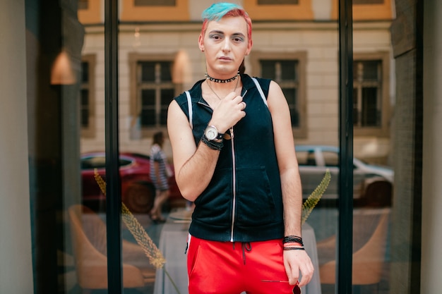 Homoxesual man avec maquillage et hairtsyle coloré dans des vêtements élégants, posant en plein air devant la fenêtre sur la rue.