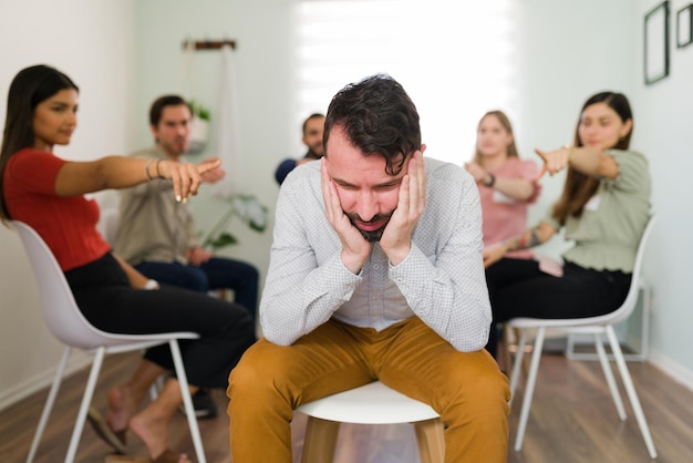 Honte et culpabilité. Groupe de personnes en cercle pointant vers un homme latin triste. Jeune homme déprimé se sentant jugé pour ses problèmes et ses dépendances