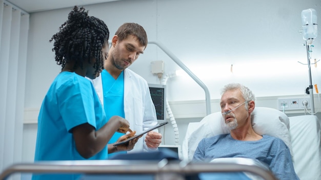 Photo À l'hôpital, un homme âgé couché sur le lit parle au médecin et à l'infirmière qui utilisent la technologie des tablettes pour guérir les patients