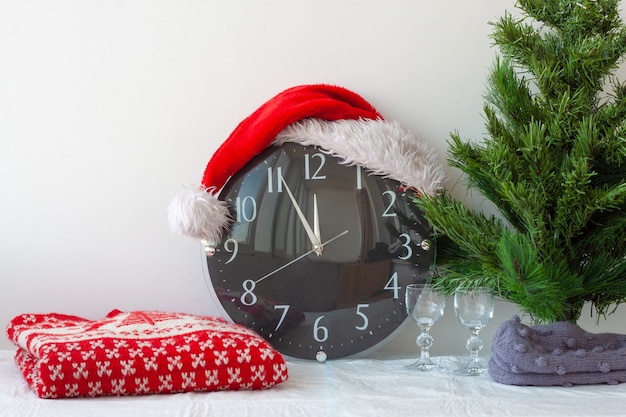 Une horloge dans un chapeau de père noël à côté d'un arbre de noël artificiel et d'un pull de noël