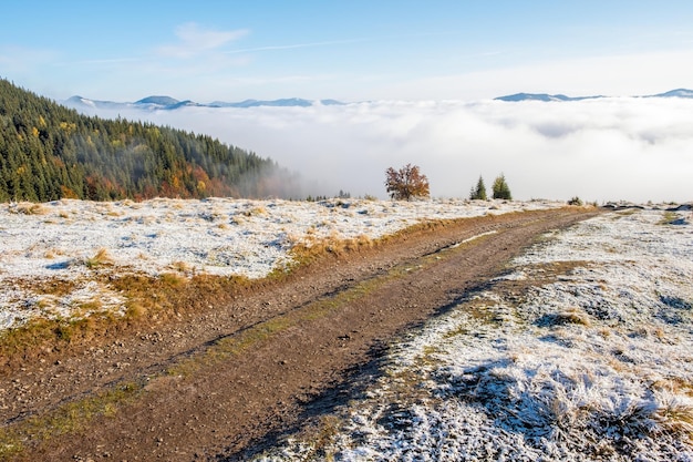 Hors route rurale du sommet aux montagnes dans les nuages au lever du soleil en automne avec la première neige Beau paysage avec le ciel forestier des Carpates ukrainiennes