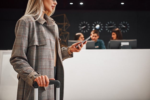 Hôtel personnel en attente d'un client. Portrait complet d'une femme d'affaires souriante et prospère tirant une valise et parlant au téléphone à l'hôtel. Gros plan de femme blonde avec smartphone