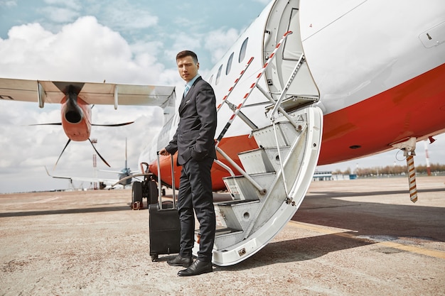 Hôtesse de l'air debout sur la piste près d'un jet d'avion. Avion de passagers moderne. Un homme multiracial confiant avec des bagages porte un uniforme et regarde la caméra. Aviation civile commerciale. Concept de voyage aérien