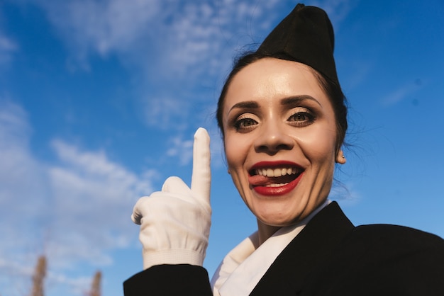 L'hôtesse de l'air drôle de belle jeune femme en uniforme montre la langue, sur fond de ciel bleu