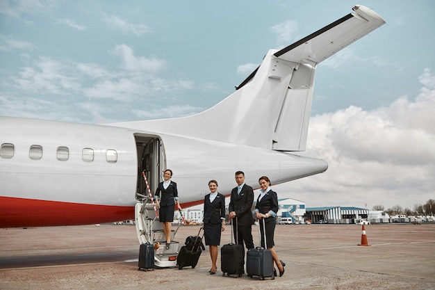 Hôtesse de l'air et hôtesses de l'air sur la piste et l'échelle près du jet d'avion. Avion de passagers moderne. L'homme et les femmes avec des bagages portent l'uniforme. Travail en équipe. Aviation civile commerciale. Concept de voyage aérien