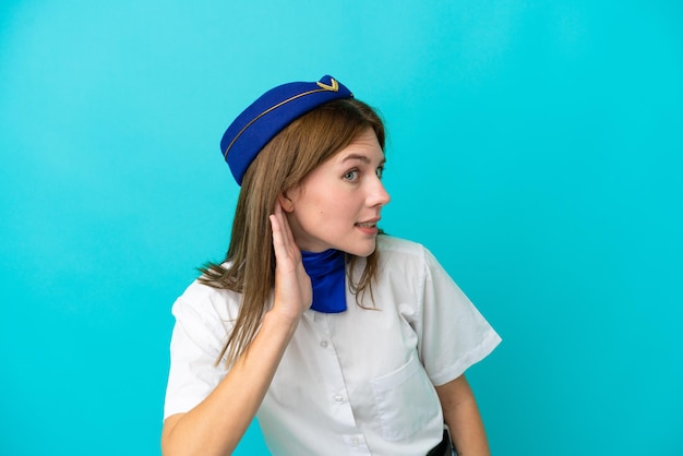 Hôtesse de l'avion anglaise femme isolée sur fond bleu écoutant quelque chose en mettant la main sur l'oreille