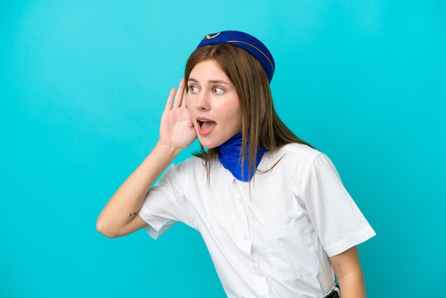 Hôtesse de l'avion anglaise femme isolée sur fond bleu écoutant quelque chose en mettant la main sur l'oreille