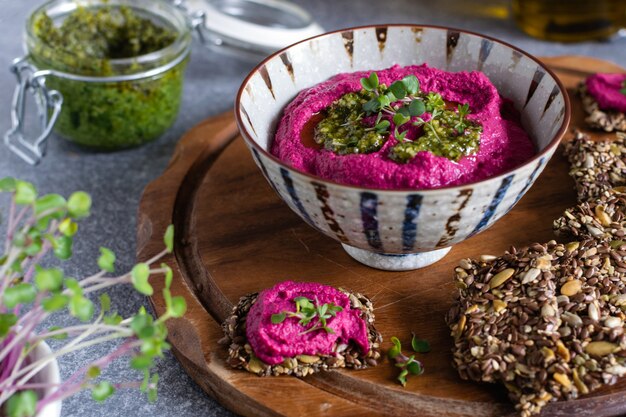 Houmous de betterave rôtie dans un bol en céramique, craquelins de graines de lin sur une planche à découper en bois, pot avec sauce pesto