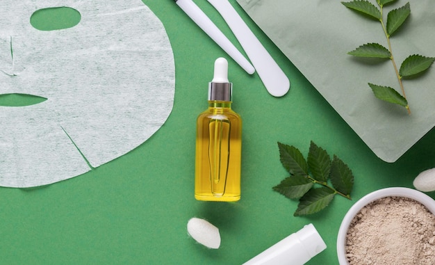 L'huile hydrophile de sérum et le masque facial cosmétique d'argile de tissu ont placé la brosse de spatule de cosmétiques, la crème hydratante. Soins de spa de beauté pour les soins du visage, cosmétologie sur fond vert avec des feuilles.