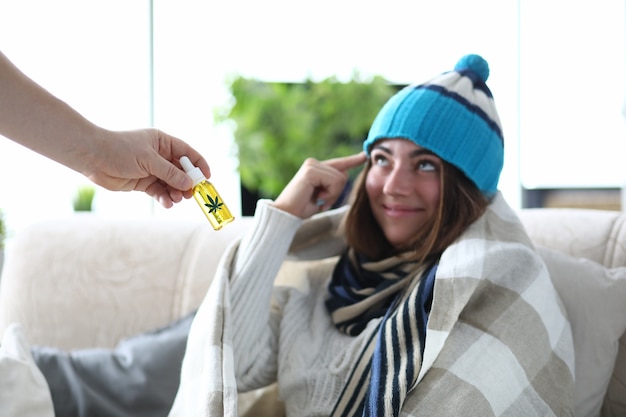 L'huile de marijuana en main libre avec une femme malade en chapeau et écharpe sous couverture