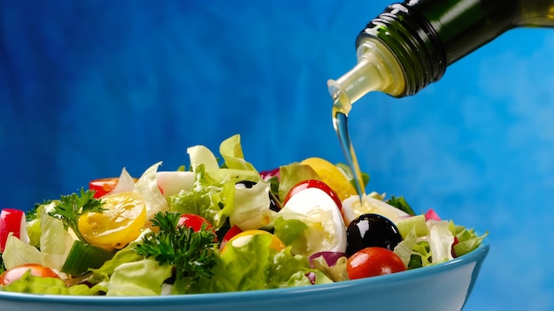 L'huile d'olive versant de la bouteille sur la salade de légumes, des aliments sains et diététiques à partir d'ingrédients frais dans un bol sur fond bleu