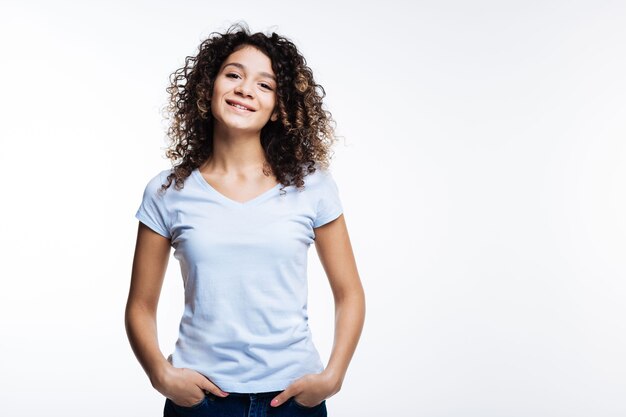 Humeur détendue. Agréable femme aux cheveux bouclés tenant ses mains dans les poches de son jean et souriant sur fond gris