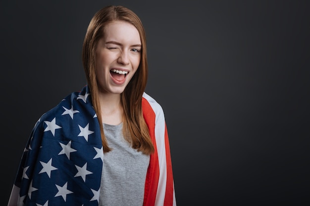 Humeur ludique. Cool fille fière créative faisant une séance photo patriotique et posant avec un drapeau en position debout isolé sur fond gris