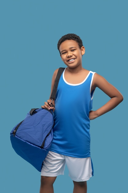 Humeur optimiste. Heureux garçon d'âge scolaire à la peau foncée en vêtements de sport avec sac bleu debout sur une photo de studio