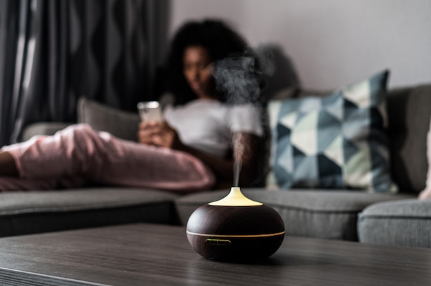 Humidificateur d'air intelligent émettant de la vapeur d'eau sur la table près d'un canapé avec une femme noire à la maison