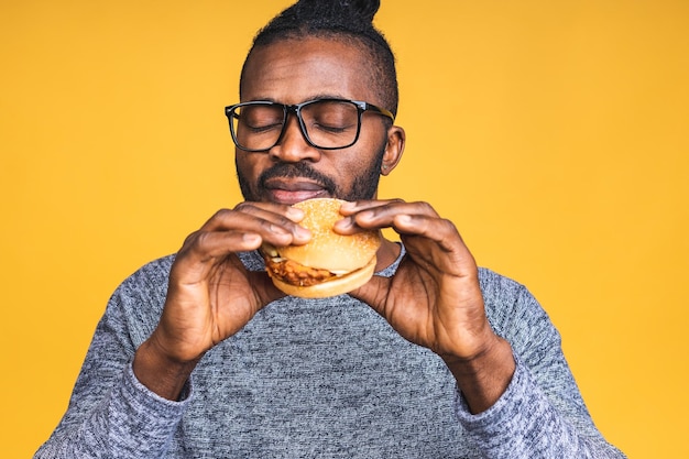 Hungry young african american man eating hamburger isolé sur fond jaune. Notion de régime.