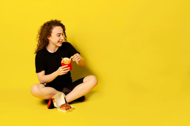 Hungry Young Curly Woman Eating French-Fries On Yellow Studio Background. Concept de repas de triche. Espace libre