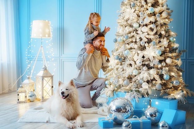 Un husky blanc se trouve près du sapin de Noël pendant que papa soulève sa fille pour l'aider à décorer le sapin de Noël