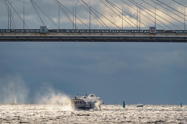 Un hydroglisseur à passagers à grande vitesse passe sous un pont à haubans par temps ensoleillé de sombres nuages de tempête en arrière-plan