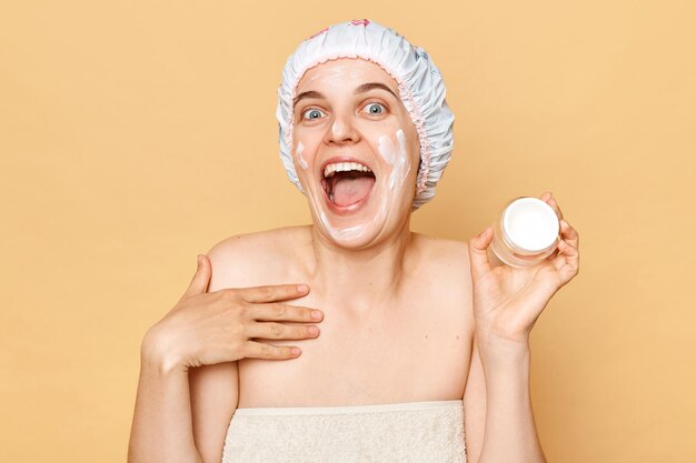 Photo hygiène et dermatologie soins de la peau sains détente et bien-être femme étonnée excitée enveloppant dans une serviette avec un bonnet de douche sur la crème de tête dans ses mains isolées sur fond beige