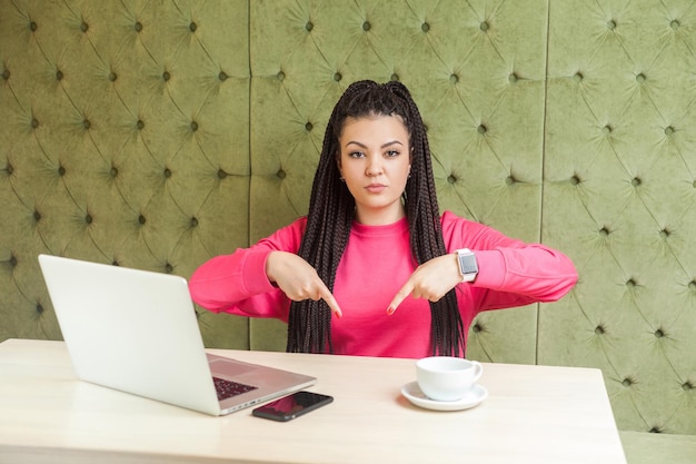 Ici et maintenant. Portrait d'une jeune femme malheureuse en colère avec une coiffure de dreadlocks noirs en chemisier rose, assise et de mauvaise humeur montrant le doigt vers le bas avec un visage bouleversé, regardant la caméra