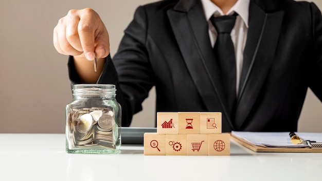 Une icône d'entreprise sur le cube en bois et la main mettant la pièce de monnaie dans la finance de concept d'argent de bouteille d'épargne