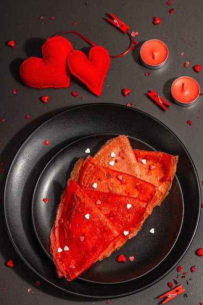 Idée de petit-déjeuner romantique, crêpes rouges ou crêpes fines garnies de cœurs sucrés. Nourriture festive pour la Saint-Valentin ou la fête des mères. Lumière dure à la mode, ombre sombre. Fond de béton de pierre noire, vue de dessus