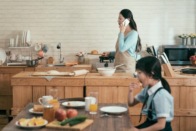 ignorer la dépendance aux gadgets familiaux. femme asiatique occupée mère tenant une assiette en bois avec croissant marchant dans une cuisine moderne parlant avec son mari sur téléphone portable le matin. petite fille mangeant prenant son petit déjeuner