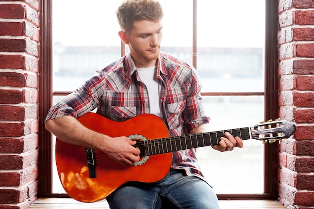 Il a une âme créative. Beau jeune homme jouant de la guitare acoustique alors qu'il était assis sur le rebord de la fenêtre