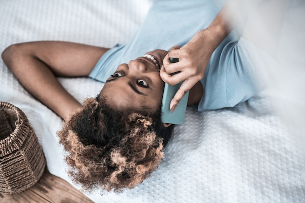 Il est temps de discuter. Sourire mignon jeune femme à la peau sombre, parler sur smartphone allongé sur le lit de bonne humeur