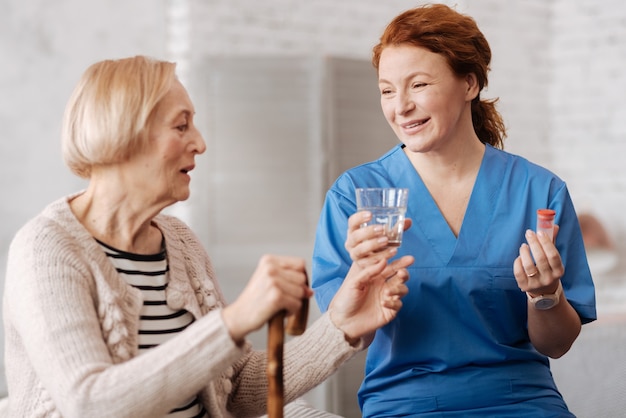 Il est temps d'être en bonne santé. Enthousiaste femme habile concentrée donnant à son patient un verre d'eau pour prendre ses pilules tout en prenant soin de la santé des vieilles dames