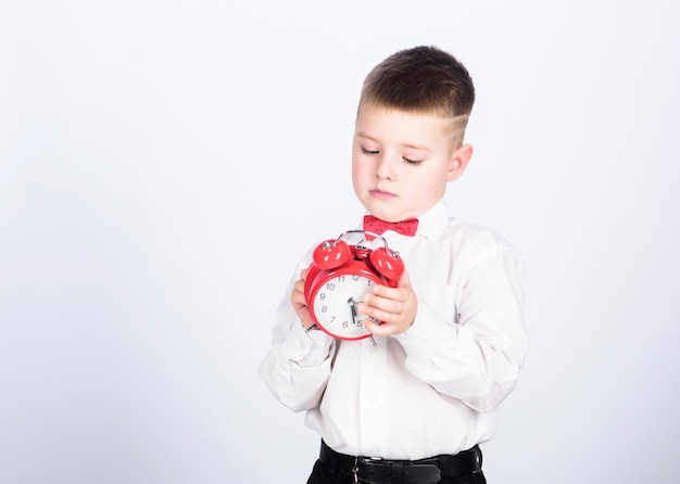 Il est temps Horaire et timing Routine du matin Écolier avec réveil Enfant adorable garçon chemise blanche noeud papillon rouge Développer l'autodiscipline Mettre en place un réveil Enfant petit garçon tenir une horloge rouge