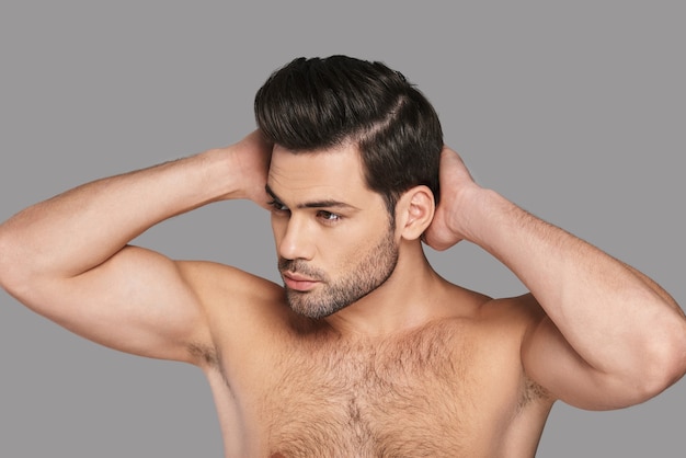 Il est totalement charmant. Beau jeune homme regardant loin et gardant les mains derrière la tête en se tenant debout sur fond gris