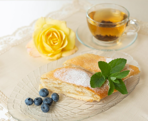Il y a deux tranches de tarte aux pommes myrtilles et feuilles de menthe sur l'assiette une tasse de thé à côté et la table est décorée d'une rose vivante