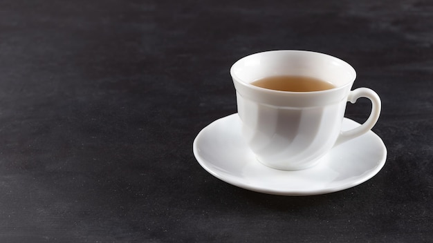 Il y a une tasse blanche avec du thé sur la table. Sur fond noir.