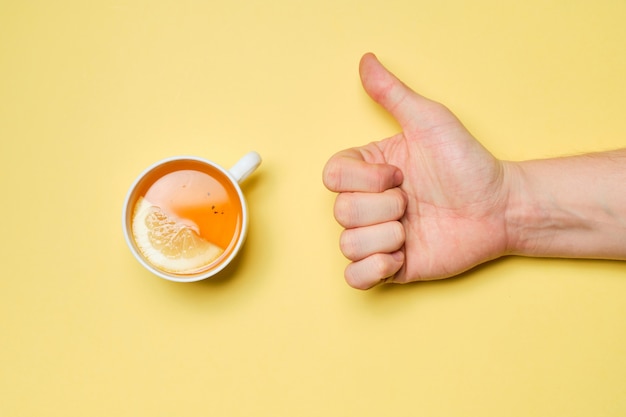 Photo l'image abrégée est appréciée par la main à côté d'une tasse de thé au citron sur fond jaune. vue de dessus.