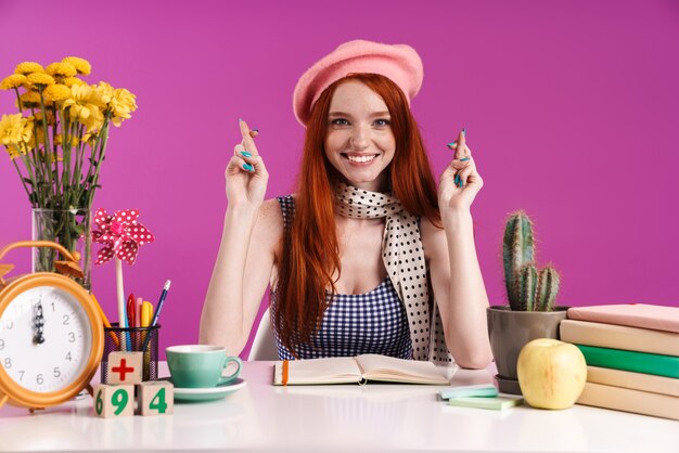 Image d'une adolescente excitée gardant les doigts croisés tout en étudiant avec des cahiers d'exercices isolés sur un mur violet