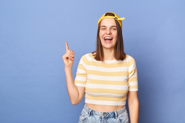 Image d'une adolescente heureuse excitée étonnée portant un t-shirt rayé et une casquette de baseball pointant vers l'espace publicitaire pour la promotion posant isolé sur fond bleu