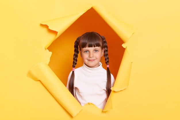 Image d'adorable charmante petite fille satisfaite avec des tresses portant un col roulé blanc posant dans un trou déchiré de mur de papier jaune regardant la caméra exprimant des émotions calmes