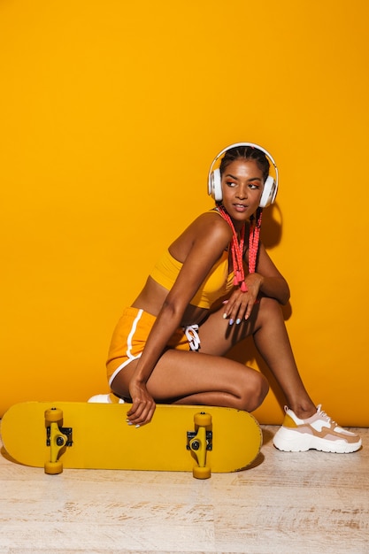 Image d'une adorable femme afro-américaine écoutant de la musique avec des écouteurs alors qu'elle était assise sur une planche à roulettes isolée sur un mur jaune