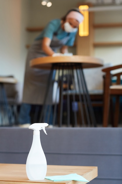 Photo image d'arrière-plan verticale d'un vaporisateur désinfectant sur une table dans un café avec un nettoyage d'une travailleuse méconnaissable, espace de copie