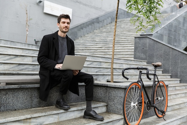 Image de beau jeune homme d'affaires assis à l'extérieur à l'aide d'un ordinateur portable.