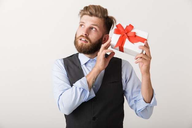 Image d'un beau jeune homme barbu debout isolé sur un mur blanc tenant une boîte cadeau.