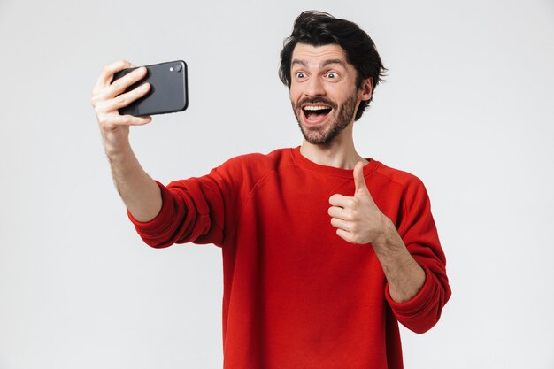 Image d'un beau jeune homme excité posant sur un mur blanc prendre un selfie par téléphone mobile montrant les pouces vers le haut.