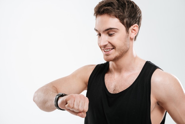 Image d'un beau jeune homme gai vêtu d'un t-shirt noir debout sur un mur blanc en regardant la montre.