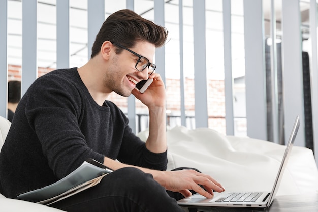 Image d'un beau jeune homme heureux à la maison à l'intérieur à l'aide d'un ordinateur portable parlant par téléphone mobile.