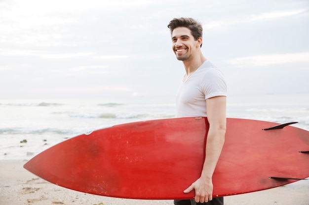 Image d'un beau jeune homme joyeux et joyeux surfeur avec surf sur une plage à l'extérieur.