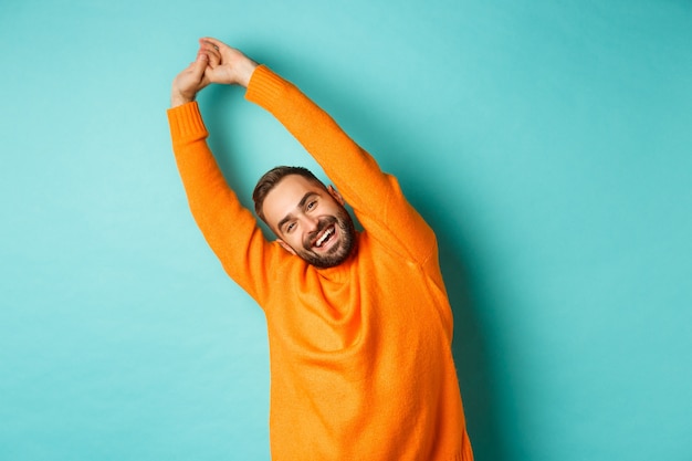 Image de beau jeune homme s'étendant les mains et souriant après un bon repos, debout en pull orange sur fond bleu clair.