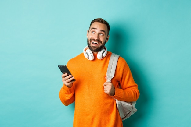 Image de beau jeune homme avec sac à dos et écouteurs, imagerie ou réflexion tout en utilisant un smartphone, debout sur fond bleu clair.